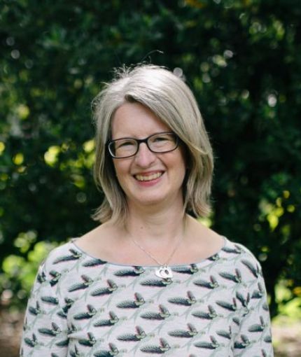 Photo of a woman standing in front of dark green leaves, wearing a green and cream top and glasses,smiling