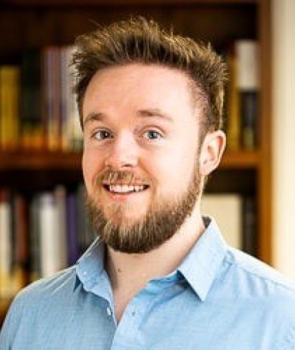 Professional headshot against a bookshelf background. Light blue shirt with a short beard.