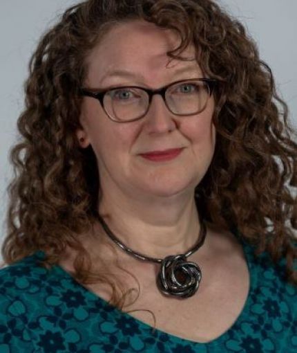 Woman with long red curly hair and glasses looking to camera.