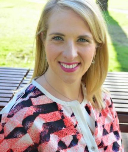 Katie has long blonde hair and fair skin. She's wearing a colorful blouse 