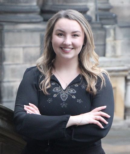 a smiling woman dressed in a black blouse standing with arms crossed outside of Old College
