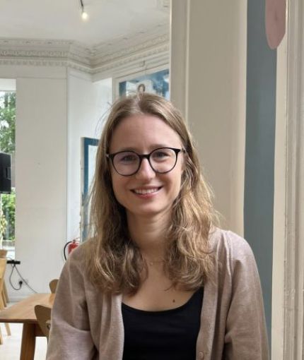 A photo of Alina, wearing a black top and beige cardigan, smiling at the camera