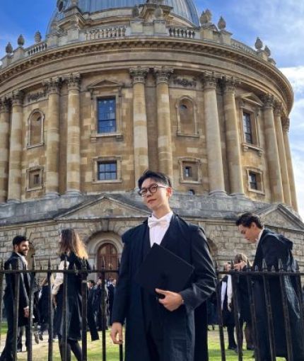 Outdoor portrait of Taehwan Kim in academic attire, standing in front of a historic library building.