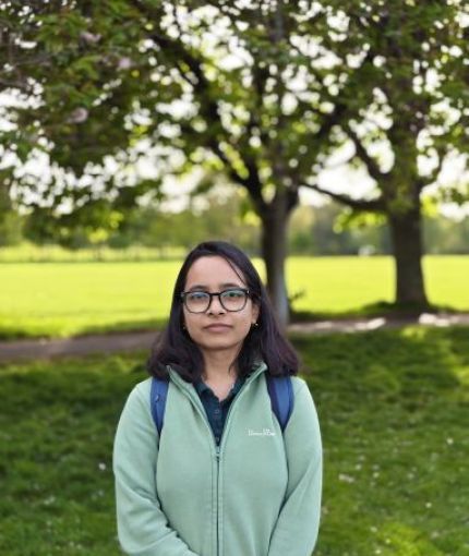 Image of Nabanita wearing a dark green t-shirt, a light green jumper, and a spectacle. She is standing at the Meadows, while some of the green stretches and trees visible around her. 