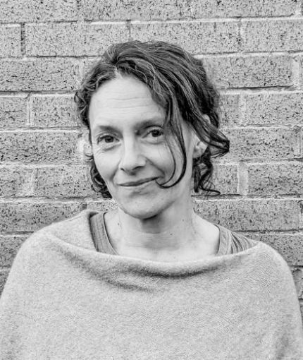 Shows a middle-aged woman turned to one side with dark hair against a brick wall