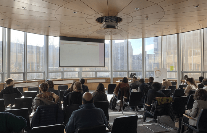 Photo taken from the back of the conference room, with people seated and looking at a projector screen