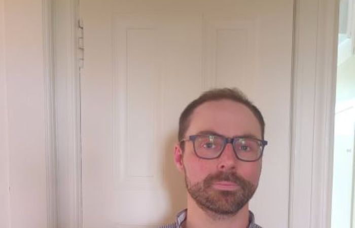 A picture of Gilbert Ramsay wearing a checked shirt and standing by a door.