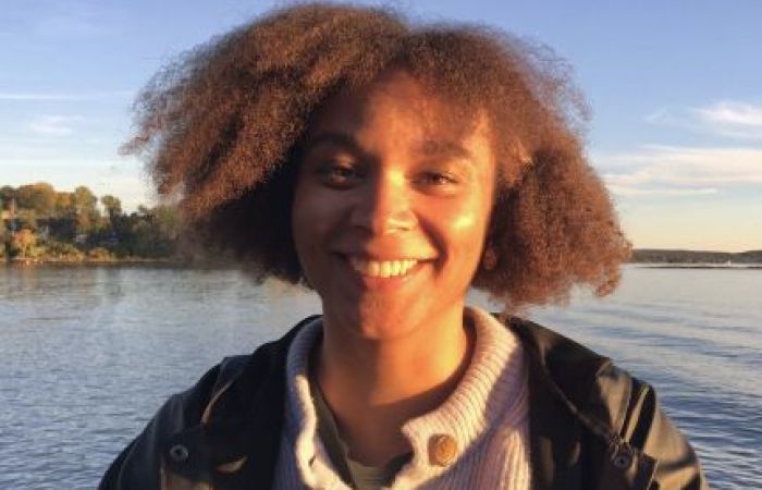 A photo of me outdoors. I am wearing a light blue cardigan and a black rain jacket. My hair is open and I am smiling at the camera.