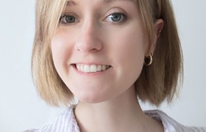 Smiling woman with chin length, blonde hair, wearing a white and purple striped shirt and gold earrings.