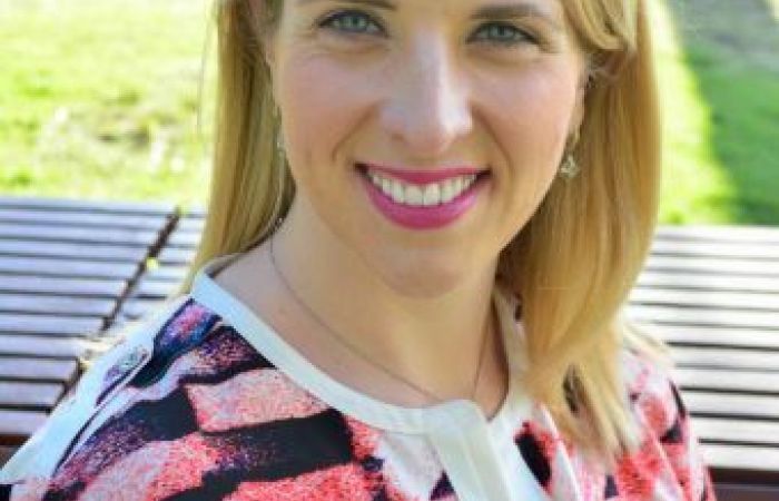 Katie has long blonde hair and fair skin. She's wearing a colorful blouse 