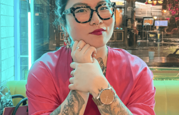 a tattooed Korean woman with glasses sitting at a table backlit with blue neon lights