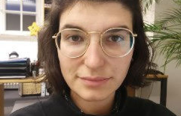 Lara's face and upper torso. They wear a black jumper, glasses and their hair is in a bun.