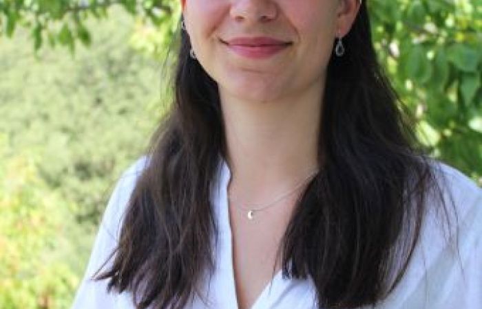 The image is a headshot of Hadewych Honné, posing and smiling in front of a background of green leaves